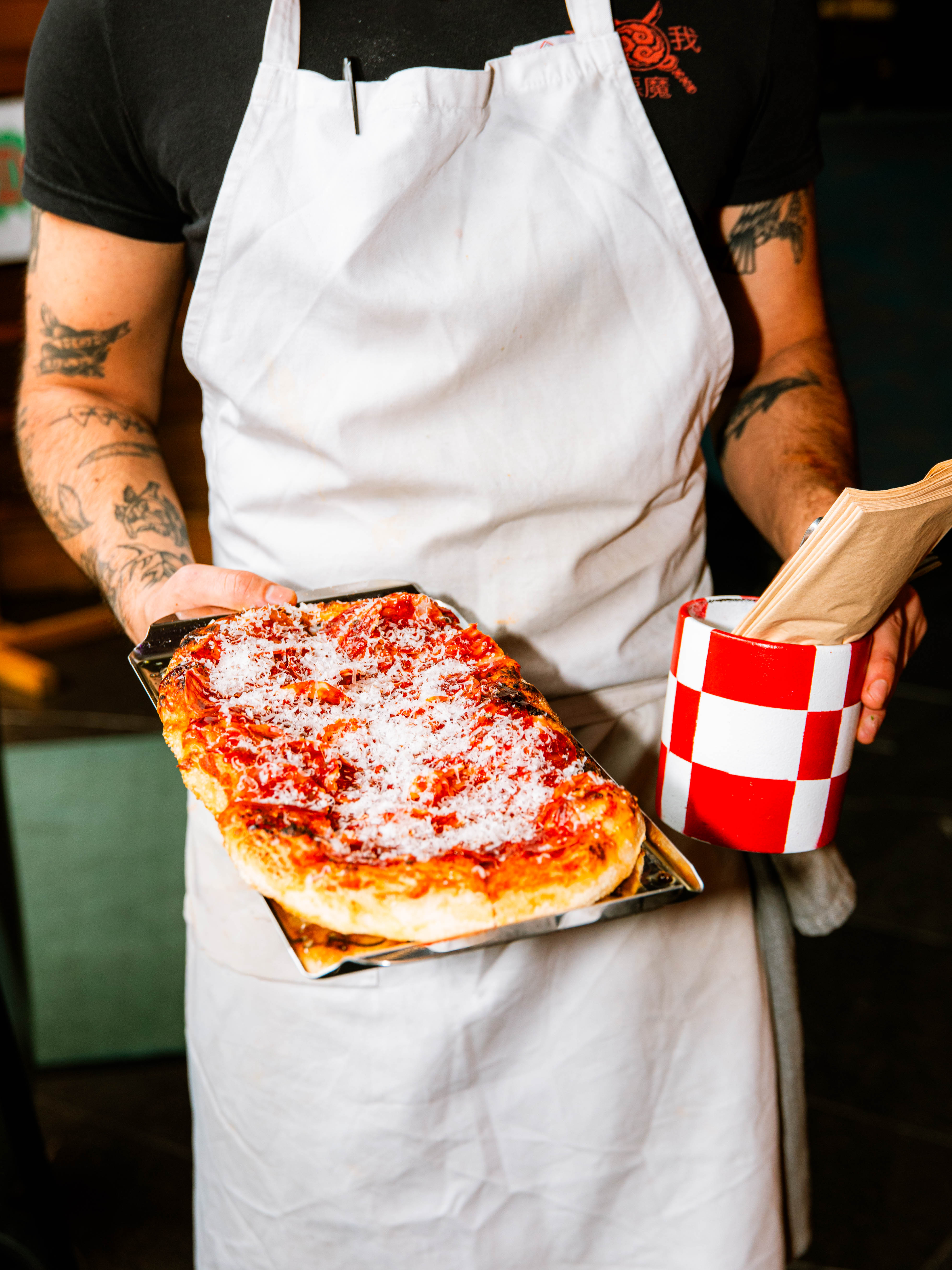 A chef at Hobo Hotel in central Stockholm holding a freshly baked tomato-based pizza with cheese in one hand and a red-and-white checkered cutlery holder in the other, ready to serve a guest.