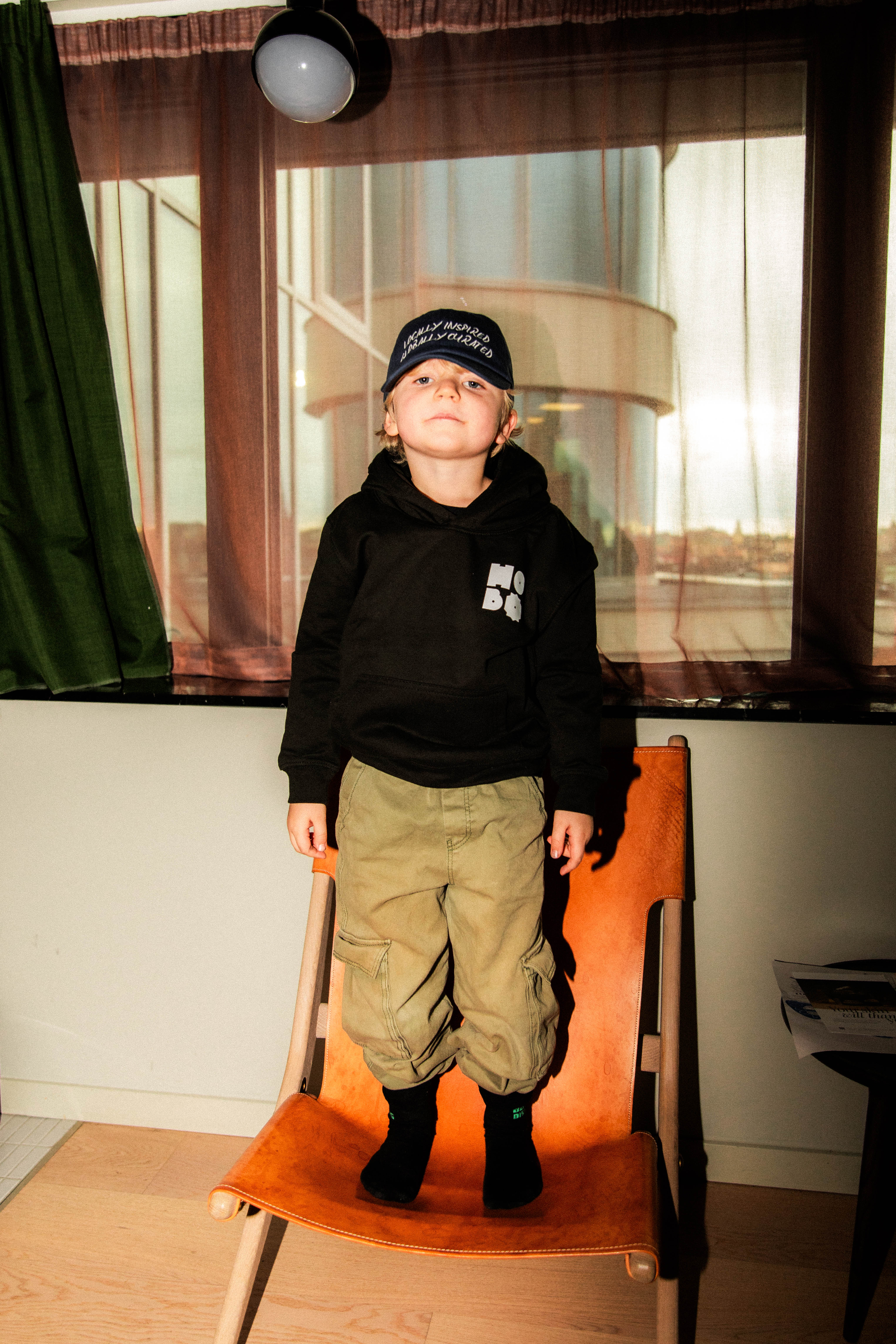 A small child standing on a leather chair looking at the camera, wearing a black Hobo merch hoodie and cap at Hobo Hotel in central Stockholm.