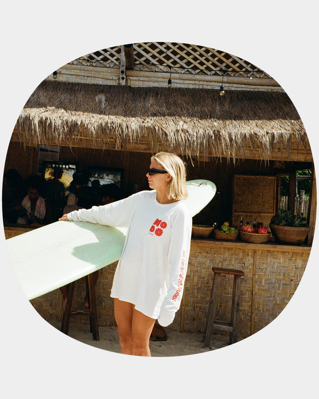 Blonde girl on a beach holding a surfboard, wearing a white long-sleeve Hobo Hotel merch shirt.