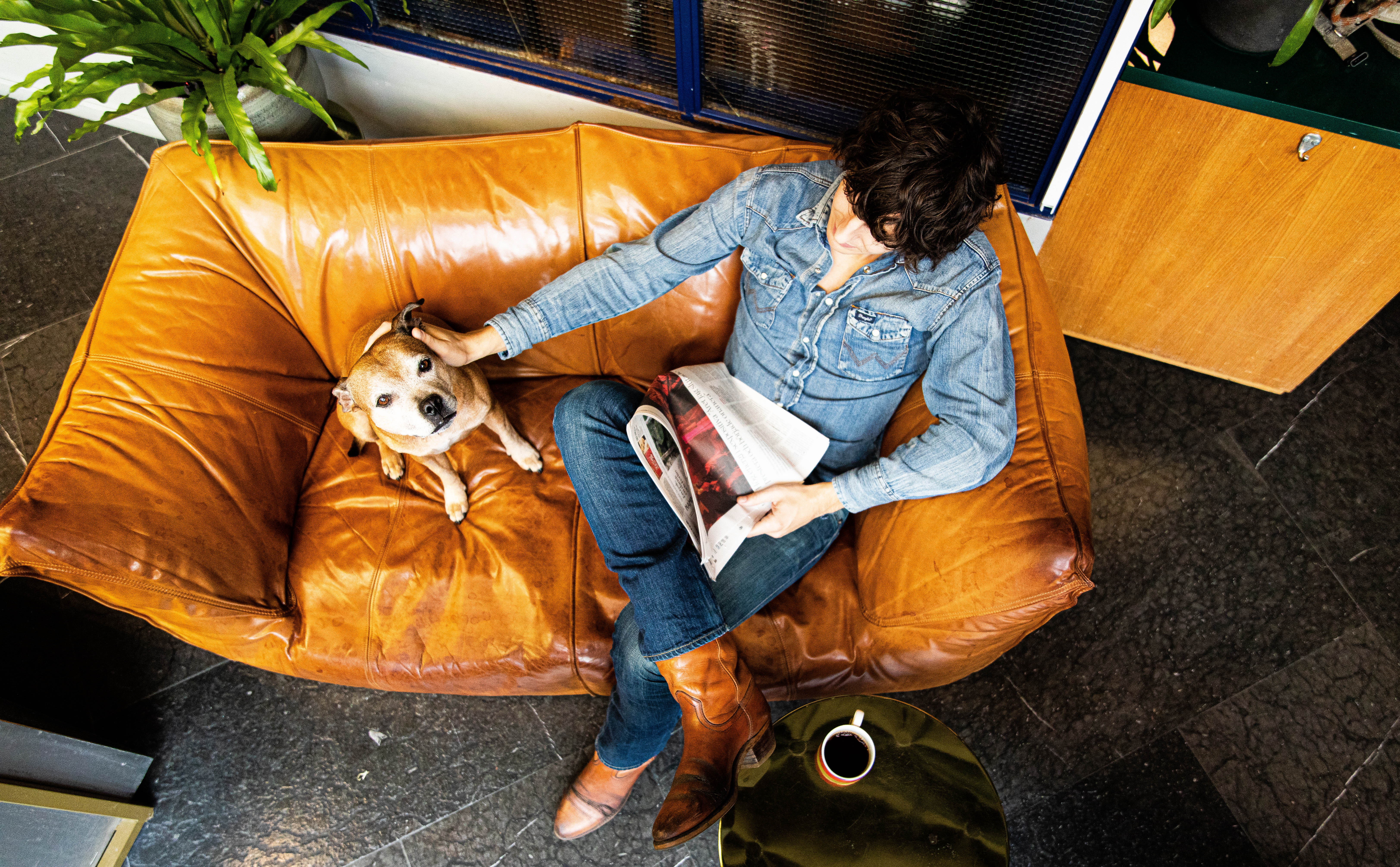 Guest in jeans and boots sitting on a leather sofa with a dog, reading a newspaper and drinking coffee in the lobby bar at Hobo Hotel in central Stockholm.